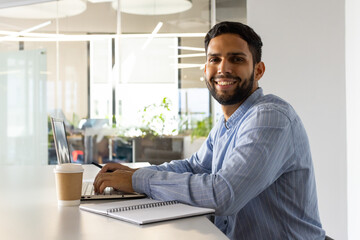 Obraz premium Man smiling and sitting at desk in office with laptop, pen and coffee cup, copy space