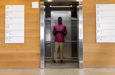 Mid-adult African American man standing in office elevator lobby holding tablet, with floor signage