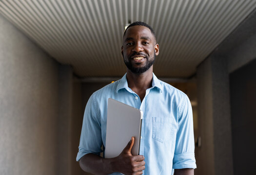 African American man standing in modern office corridor, holding silver laptop and smiling warmly