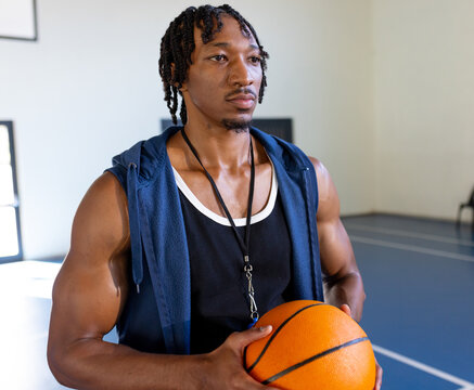 African American man holding basketball and whistle on indoor blue court, displaying focus