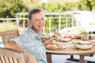 Senior woman enjoying lunch outdoors on wooden patio table with sandwiches salad bowl and rosé wine