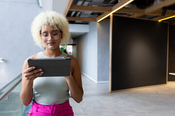Woman standing in modern office corridor leaning against glass railing, holding tablet and reading