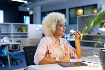 African American woman speaking into smartphone at desk in office, with laptop and tablet