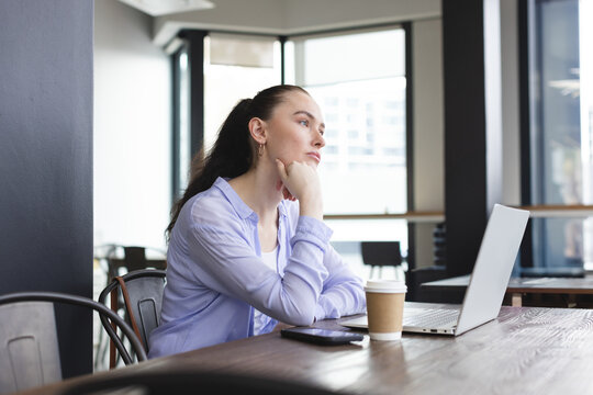 Woman leaning on hand and gazing out window at modern cafe, with laptop coffee cup smartphone - Powered by Adobe