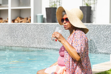 Senior woman sitting at poolside, wearing straw hat, red sunglasses, sipping from glass, copy space