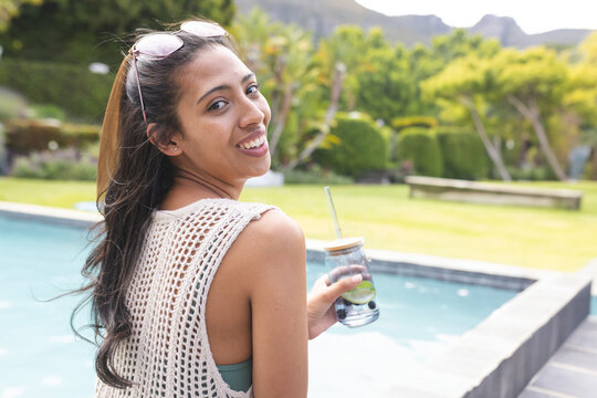 Multiracial woman sitting poolside backyard holding mason jar with lime water, smiling, copy space - Powered by Adobe