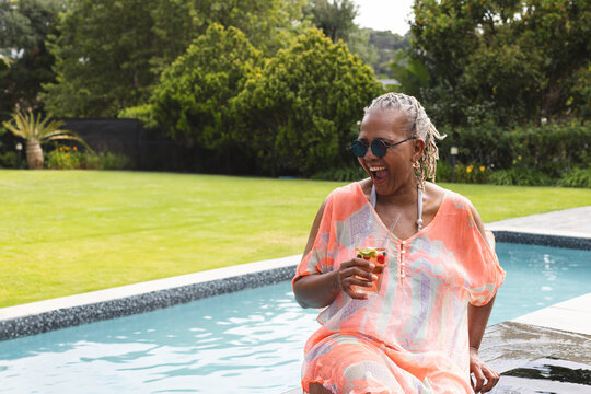 Senior African American woman sitting on backyard pool edge laughing and holding drink, copy space - Powered by Adobe