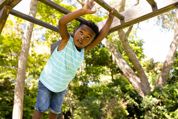 African American boy hanging from wooden monkey bars in park with tall leafy trees all around