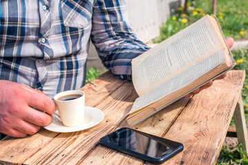  man reading book outdoors, next to wooden table and coffe cup