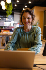 Woman in early 40s typing on laptop in open-plan office, with plants and lights