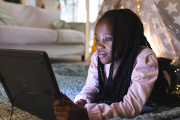 African American girl lying on stomach using tablet at home, star tent, string lights, copy space