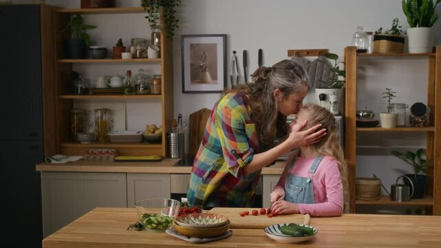 Medium full shot of happy loving Caucasian mother standing in kitchen of cozy family home, watching young daughter slice fresh vegetables for salad, then hugging and kissing girl on forehead