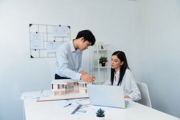 Naklejka premium Professional male asian architect using ruler to measure house model length while young beautiful caucasian colleague using laptop to analyzed data on meeting table with house model. Immaculate.
