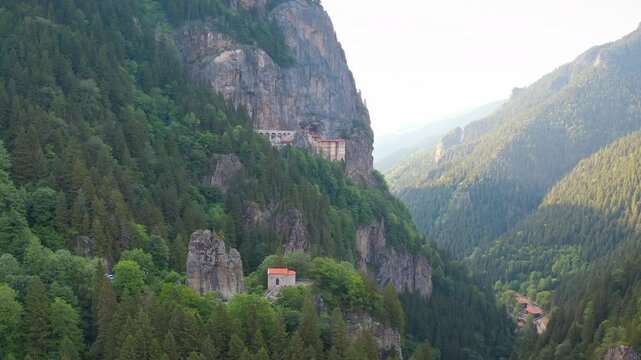 A drone shot with a fascinating turning motion approaches the historical Sumela Monastery, located on steep cliffs rising into the sky, among mountains covered with lush forests.