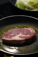 A ribeye steak is cooking in a skillet with olive oil, releasing savory aromas in a warm, inviting kitchen. Fresh vegetables are visible in the background, enhancing the culinary atmosphere.