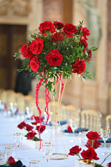 Close-up of luxurious red rose floral centerpiece on elegant wedding table with gold stand, white linen, and glassware, set in a romantic historic palace for upscale event decor inspiration

