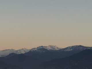 Fototapeta premium Snow-covered mountain peaks at sunset with soft gradient sky and distant layers of dark hills in the foreground. A faint airplane contrail is visible.