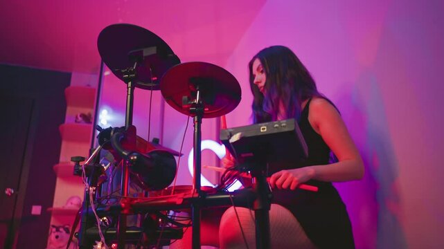 Upward angle showing girl practicing electronic drums under pink and purple studio lights, seated with focused expression, mirror reflecting glowing heart light and background