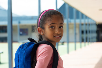 Girl child standing in covered school corridor, wearing pink headband and carrying blue backpack