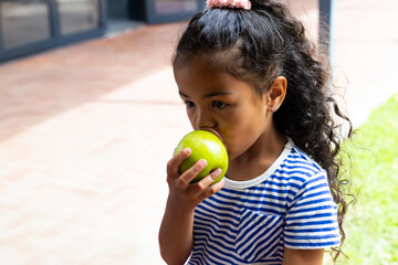 Multiracial girl child biting bright green apple on brick patio at home, sunny snack moment