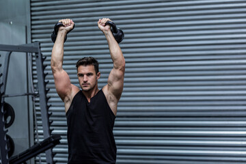 Man lifting kettlebells overhead at industrial-style gym, with weight rack and rubber flooring