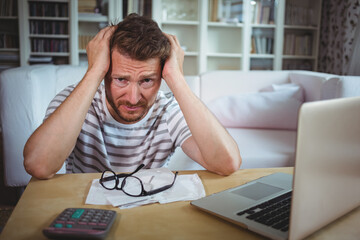 Mid-adult man leaning at table at home holding head amid papers, glasses, calculator, laptop