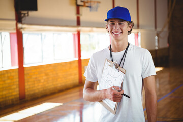 Teenage boy standing in basketball gym, holding clipboard with play diagram and whistle, copy space