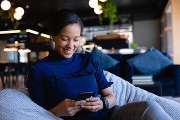 Woman sitting in cushioned lounge chair in co-working lounge, holding smartphone and smiling