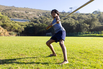 Multiracial boy standing on grassy soccer field, gripping javelin pole near soccer goals