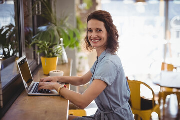 Woman typing on silver laptop at wooden cafe counter by large window, with yellow potted plant