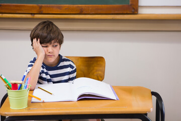 Boy leaning head on hand at desk in classroom, notebook and colored pencils in metal cup