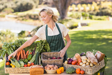 Woman arranging fresh produce on table at outdoor farmers market, with woven baskets of vegetables