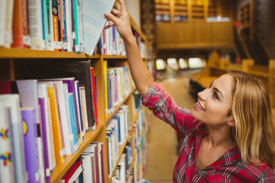 Female student reaching for hardcover book on wooden shelf in library aisle by tables, copy space