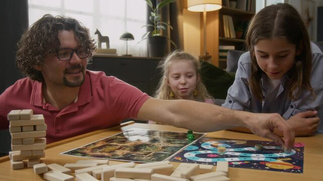 Medium shot of cheerful Caucasian father with limb difference disability playing roll and move game with wife, son and daughter at home, shaking, throwing dice, moving checker on field