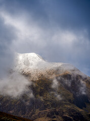 A lofty Glyderau summit materialises through drifting clouds, revealing a glistening snowcap against the wild North Eryri sky