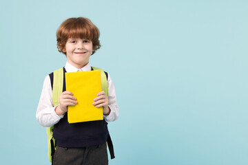 Cute schoolboy with backpack and book on light blue background, space for text