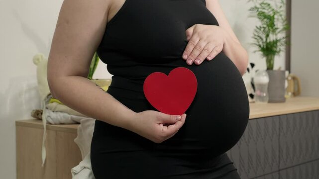 Pregnant woman holding red heart shape and feel love for unborn baby in belly. Awaiting for baby. Concept of maternity and pregnancy care
