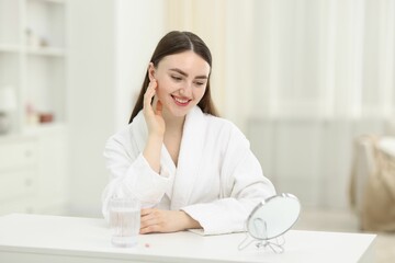 Smiling woman looking at round mirror and glass of collagen water on table indoors