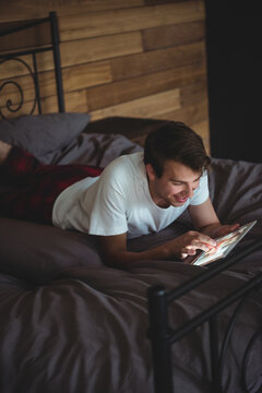 Man lying on bed on stomach tapping tablet in bedroom, featuring gray bedding, wood accent wall