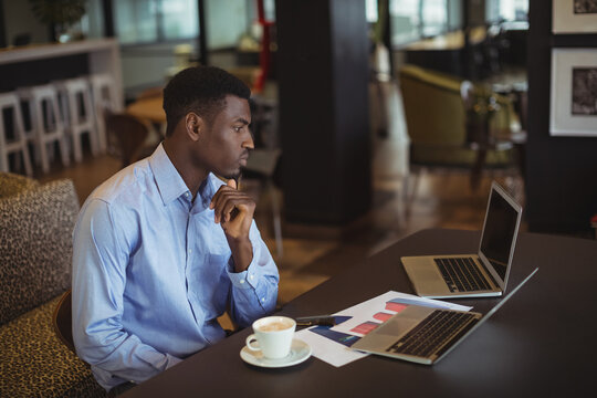 African American young man leaning forward in cafe, viewing laptops and charts, copy space