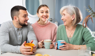 Portrait of positive young couple and senior woman sitting at table at home and drinking tea together.