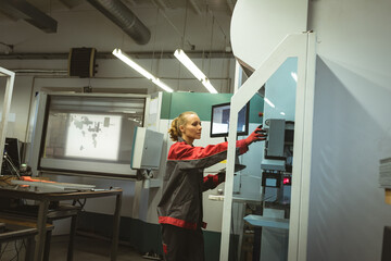 Woman adjusting controls on large industrial machine at factory workshop, wearing safety gloves