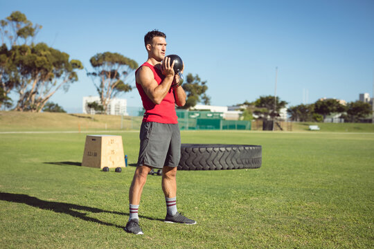 Man in his thirties holding medicine ball on training field, with plyometric box and tractor tire