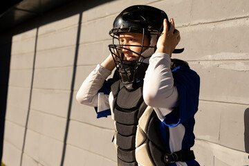 Female baseball catcher adjusting catchers helmet by concrete wall in sports dugout, copy space