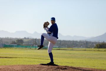 Young adult woman pitching baseball from dirt mound on grass field, with glove and blue uniform
