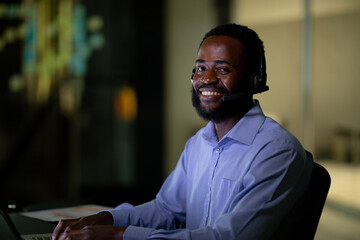 African American man smiling and typing on laptop in dimly lit office with headset and papers