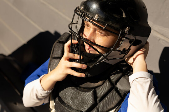 bi-racial young adult woman adjusting catcher's helmet in dugout with chest protector, blue shirt - Powered by Adobe