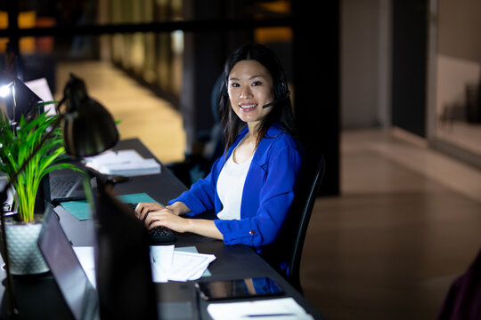 Chinese woman typing on keyboard at desk in modern open-plan office, with headset and laptop