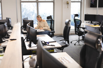 Woman writing in notebook at open-plan office desk with monitors keyboard mouse and ergonomic chair