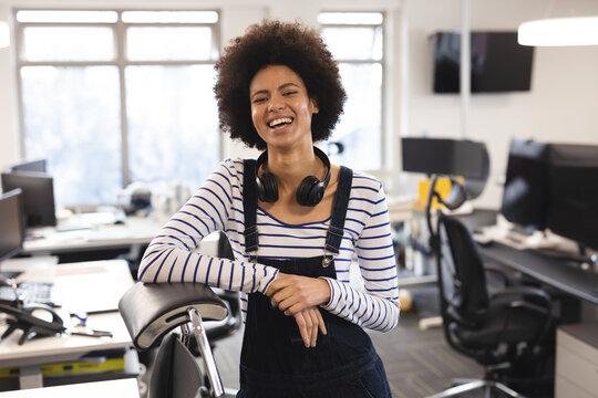 Young adult woman leaning on ergonomic office chair in open-plan office, smiling with headphones